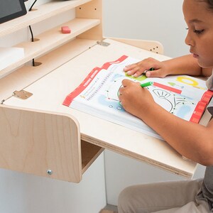 May include: A light wood wall-mounted desk with a fold-down surface. A child is sitting at the desk and drawing in a workbook with a green marker.