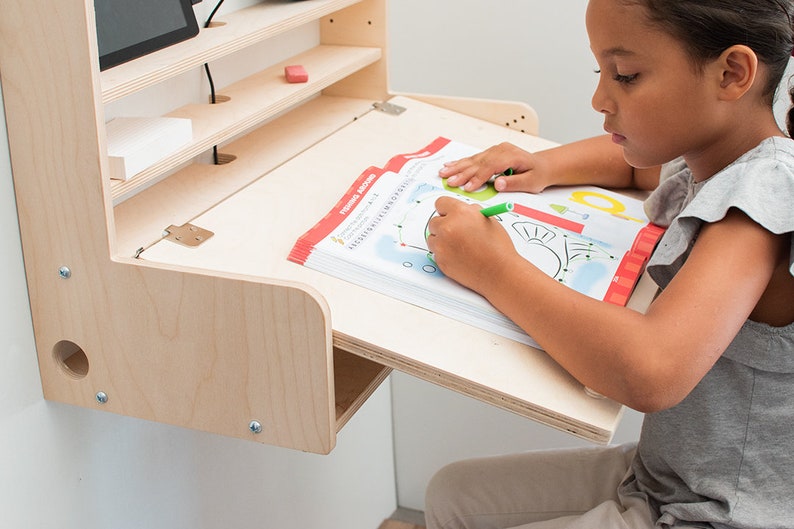 May include: A light wood wall-mounted desk with a fold-down surface. A child is sitting at the desk and drawing in a workbook with a green marker.