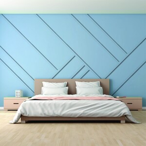 Op de afbeelding: Een wit bed met een lichtbruine hoofdeinde en een lichtroze deken. Het bed staat in een kamer met een lichtblauwe muur met een geometrisch patroon.