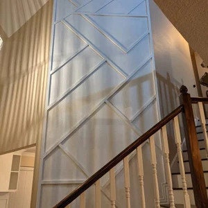 Pode incluir: Um design de painel de parede geom&eacute;trico em tons de branco e cinza claro. O painel apresenta uma s&eacute;rie de linhas e formas angulares, criando um visual moderno e art&iacute;stico. O painel &eacute; instalado em uma parede ao lado de uma escada com corrim&atilde;o de madeira.