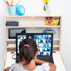 May include: A young person sits at a fold-down desk with a laptop open to a video call. The laptop screen shows a grid of people in a video conference. The desk is mounted to a wall and has a shelf above it with a blue bucket, a blue clock, and two boxes of crayons.