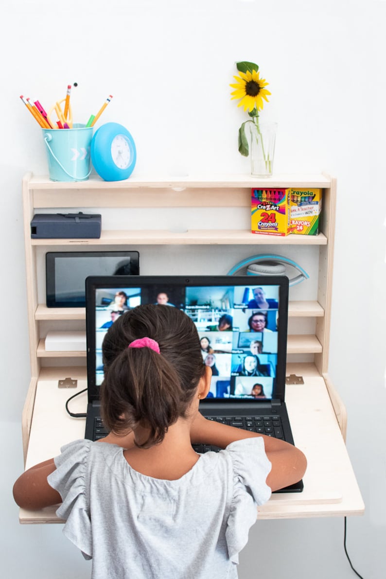 May include: A young person sits at a fold-down desk with a laptop open to a video call. The laptop screen shows a grid of people in a video conference. The desk is mounted to a wall and has a shelf above it with a blue bucket, a blue clock, and two boxes of crayons.