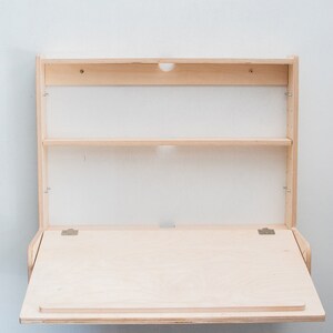 May include: A light wood folding wall desk with a shelf above the desk surface. The desk is closed and folded up against the wall.