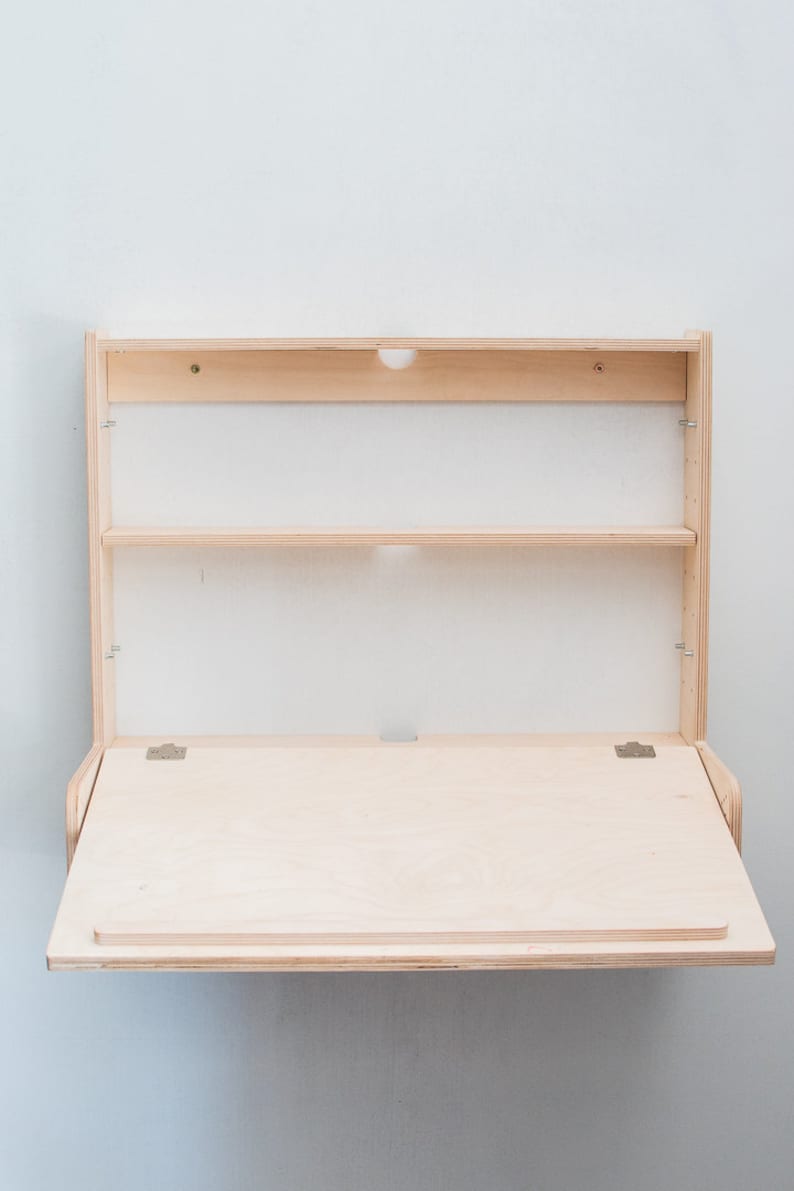 May include: A light wood folding wall desk with a shelf above the desk surface. The desk is closed and folded up against the wall.