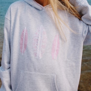 May include: A light gray hoodie with a kangaroo pocket and a drawstring hood. The front of the hoodie features three pink leaf-shaped appliqués. The appliqués have different patterns, including gingham and lace.