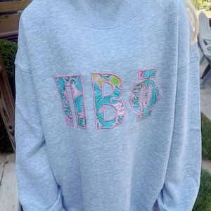 May include: A light gray mock neck sweatshirt featuring the Greek letters Pi Beta Phi embroidered on the chest. The letters are filled with a floral pattern in shades of teal, green, and pink, and are outlined with a thin pink thread border.