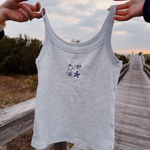 May include: A gray tank top with a simple embroidered design of a beach umbrella, two seashells, a flower, and a starfish.