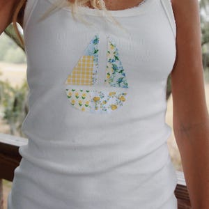 May include: White tank top with a patchwork sailboat design in yellow, blue, and white fabric.