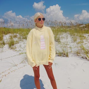 May include: A light yellow sweatshirt with a lighthouse design in pink and white. The sweatshirt is oversized and has long sleeves. The person is wearing sunglasses and standing on a sandy beach under a blue sky with clouds.