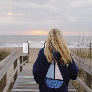 May include: A person wearing a navy blue hoodie with a white and blue sailboat design on the back. The person is walking on a wooden boardwalk towards the beach.