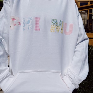 May include: White hooded sweatshirt with a pink, blue, and yellow patchwork design that spells out "PHI MU".