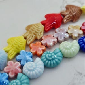 May include: A selection of brightly coloured ceramic beads in diverse shapes, such as fish, flowers, and seashells. The beads are in shades of yellow, orange, pink, blue, green, and brown. The glossy finish reflects light, creating a vibrant and playful aesthetic.