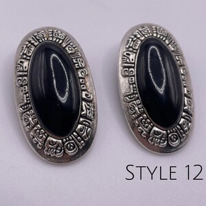 May include: A pair of silver tone earrings with black oval stones set in a detailed design. The earrings have a Mayan-inspired pattern around the stones. STYLE 12 is printed below the earrings.