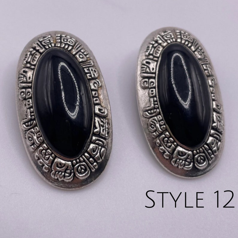 May include: A pair of silver tone earrings with black oval stones set in a detailed design. The earrings have a Mayan-inspired pattern around the stones. STYLE 12 is printed below the earrings.