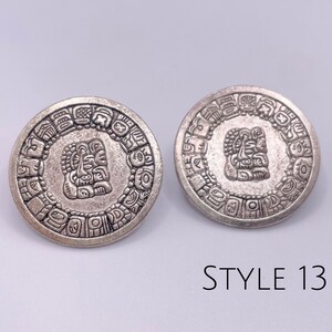 May include: A pair of silver earrings with a circular design featuring an intricate Mayan-inspired pattern. The earrings have a raised, detailed image of a face in the center. The text "STYLE 13" is visible below the earrings.