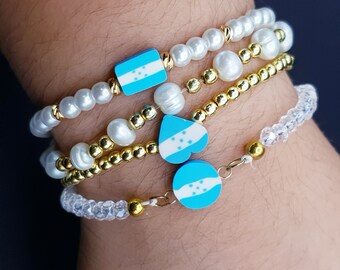 May include: A set of four bracelets featuring white pearls, gold beads, and blue and white charms with the flag of Honduras. The charms are in the shape of a rectangle and a heart.