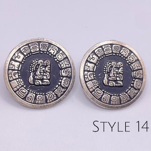May include: A pair of silver earrings with a black background and a silver Aztec-style design. The earrings are round and have a diameter of approximately 1 inch. The design features a central face surrounded by a circle of smaller designs. The earrings are labeled "STYLE 14".