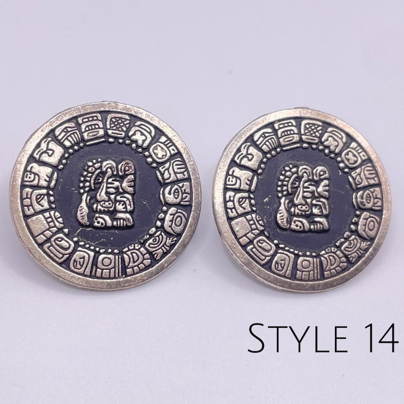 May include: A pair of silver earrings with a black background and a silver Aztec-style design. The earrings are round and have a diameter of approximately 1 inch. The design features a central face surrounded by a circle of smaller designs. The earrings are labeled "STYLE 14".
