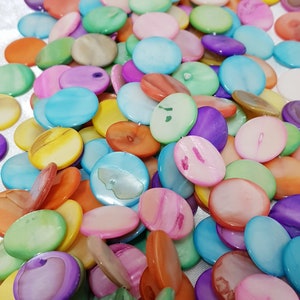 Puede incluir: Un conjunto de cuentas ovaladas planas e iridiscentes de varios colores. Las cuentas son de tonos azules, verdes, amarillos, naranjas, rosas y morados. Tienen un aspecto brillante y nacarado y están dispersas sobre la superficie.