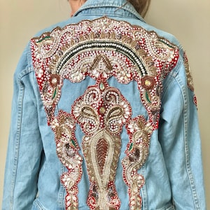 May include: Light blue denim jacket with ornate, colorful beadwork on the back. The design features red, gold, and white embellishments in a detailed, symmetrical pattern. The jacket has button closures and a classic collar.