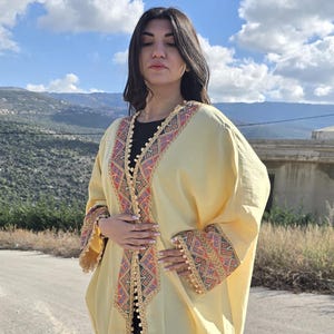May include: A long, flowing, pale yellow garment with colorful embroidered trim on the sleeves and neckline. The garment is worn over black leggings and paired with black flats. The background features a mountainous landscape under a blue sky.