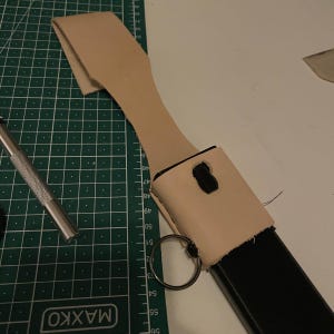 May include: A light brown leather holster with a black metal object inside. The holster has a metal ring attached to it and is sitting on a green cutting mat.