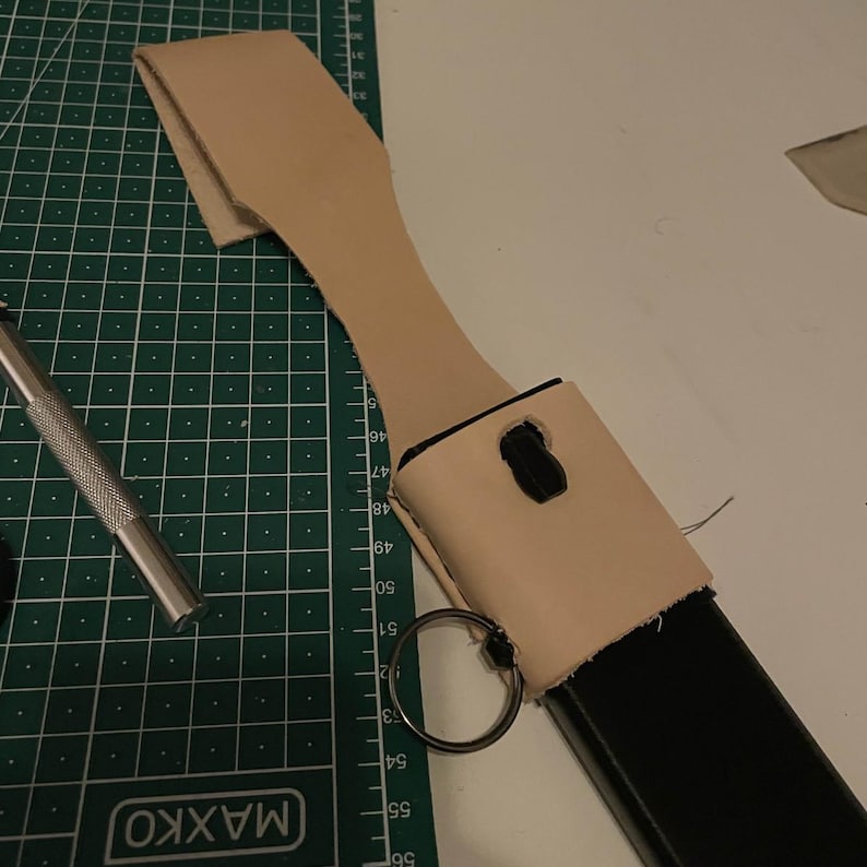 May include: A light brown leather holster with a black metal object inside. The holster has a metal ring attached to it and is sitting on a green cutting mat.