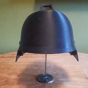 May include: A black plastic helmet with a raised design on the top. The helmet is on a metal stand.