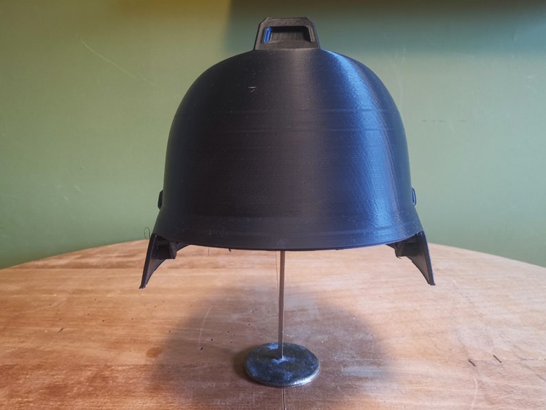 May include: A black plastic helmet with a raised design on the top. The helmet is on a metal stand.
