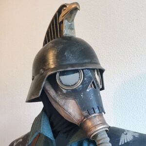 Puede incluir: Un casco de m&aacute;scara de gas negro y marr&oacute;n con un &aacute;guila dorada en la parte superior. El casco tiene una parte frontal en forma de calavera y una pieza ocular grande y redonda.
