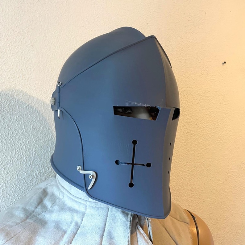 May include: A blue medieval helmet with a cross design on the front. The helmet has eye slits and is made of a sturdy material. The helmet is designed to protect the head and face.