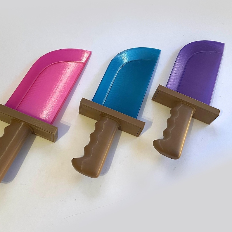 May include: Three colorful toy knives. The blades are pink, blue, and purple, with brown handles. The handles have a curved, ergonomic design. The knives are arranged in a row, with the blades facing up. The background is white.