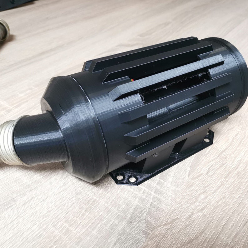 May include: A black 3D printed object with a cylindrical shape and a series of vertical slots. The object has a rounded end and a flat base with mounting holes.