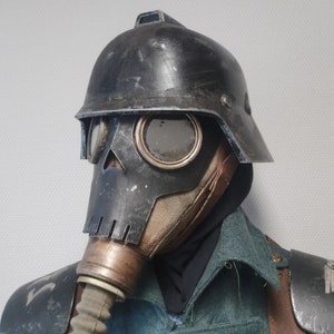 K&ouml;nnte beinhalten: Eine schwarze und braune Gasmaske mit Totenkopfdesign und einem Metallhelm. Der Helm hat ein Visier und einen Riemen.