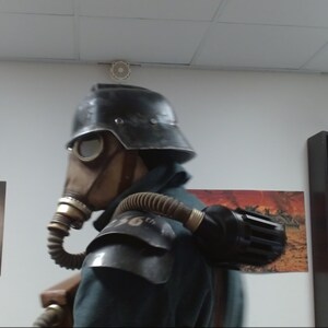 May include: A person wearing a black helmet and gas mask with a large black air tank strapped to their back. The person is wearing a dark green jacket and has a brown leather strap across their chest.