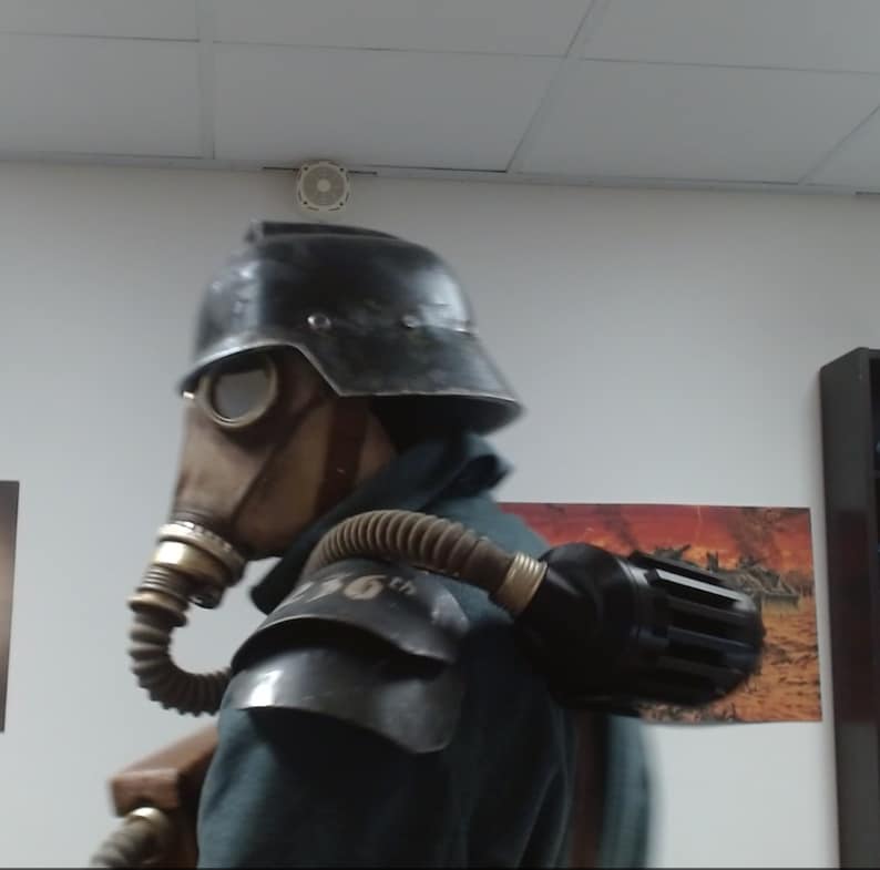 May include: A person wearing a black helmet and gas mask with a large black air tank strapped to their back. The person is wearing a dark green jacket and has a brown leather strap across their chest.