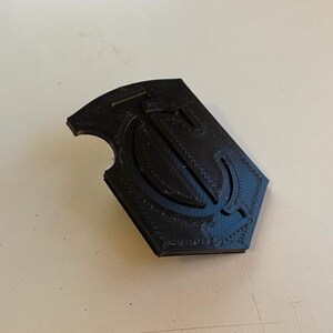 Op de afbeelding: Een zwart 3D-geprint embleem met een gestileerde letter "T" in een schildvorm.