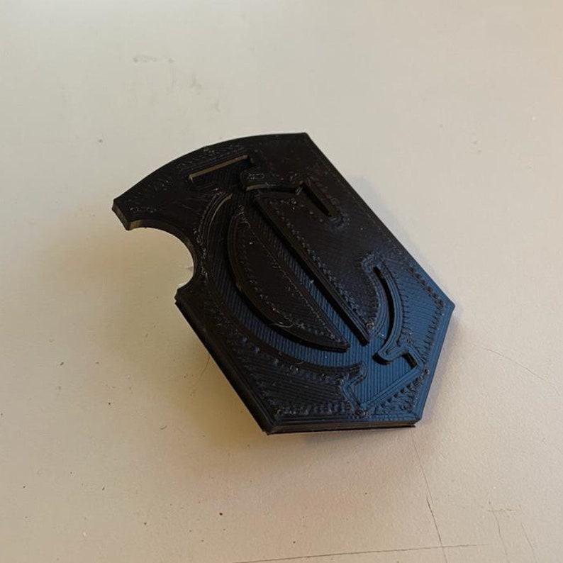 Op de afbeelding: Een zwart 3D-geprint embleem met een gestileerde letter "T" in een schildvorm.