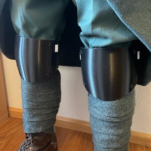 May include: A pair of black knee pads with a textured surface. The knee pads are worn over grey fabric.