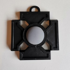 May include: A black, geometric pendant with a circular, grey centre. The pendant has a cross-like design with a loop at the top for hanging. The design is made of a textured material.