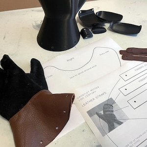 May include: A black suede glove with a brown leather cuff, a black 3D-printed gauntlet component, several small black plastic plates, brown leather straps, and printed instructional diagrams labelled Gauntlet Mitten 15th Century.