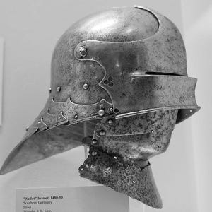 May include: A grayscale image of a steel sallet helmet from Southern Germany, dating from 1480-90. The helmet features a rounded top, a visor, and a pointed lower section. The helmet is riveted and has decorative elements.