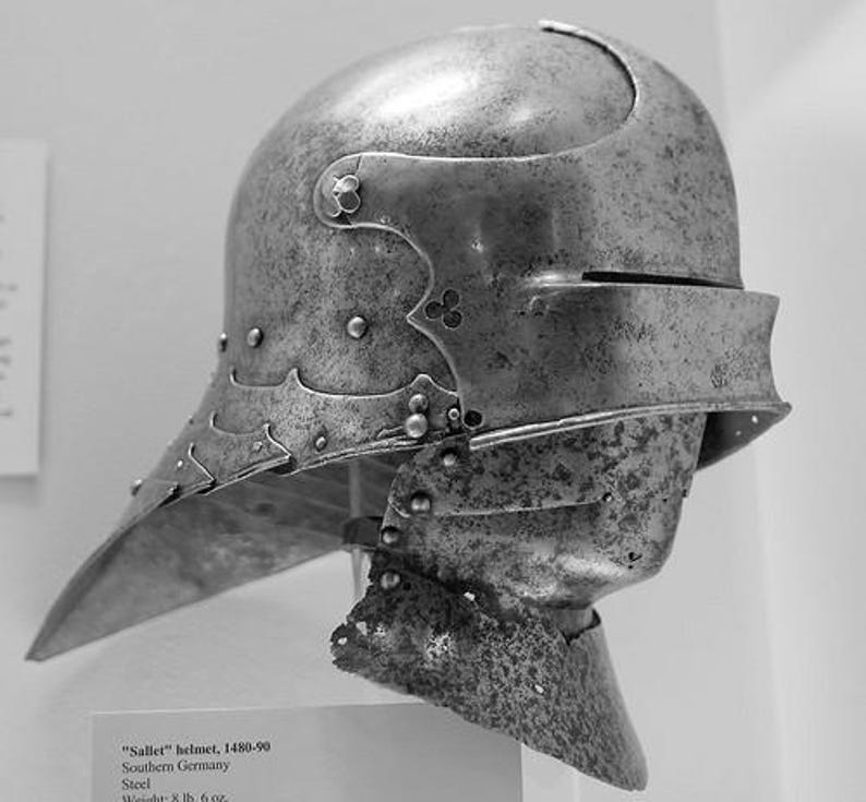 May include: A grayscale image of a steel sallet helmet from Southern Germany, dating from 1480-90. The helmet features a rounded top, a visor, and a pointed lower section. The helmet is riveted and has decorative elements.