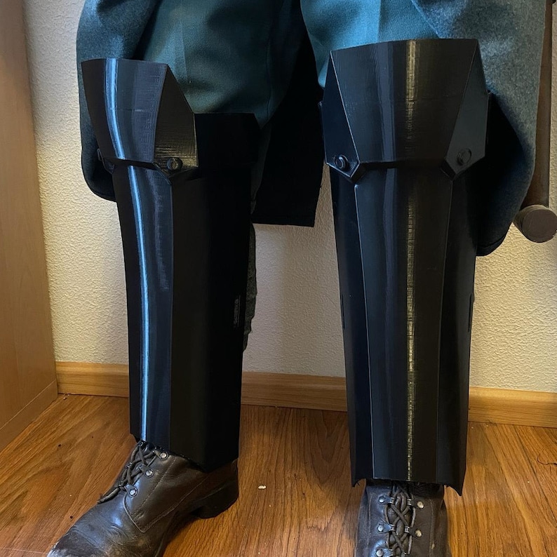 May include: A pair of black 3D printed knee pads with a geometric design. The knee pads are worn over pants and have a smooth, shiny finish.