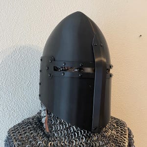 以下が含まれることがあります： 黒い中世のヘルメット。尖った上部と顔面ガードが付いています。ヘルメットはリベットで留められており、金属製です。ヘルメットをかぶっている人は、鎖かたびらも着用しています。