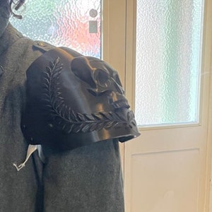 May include: A black shoulder pauldron with a skull design and laurel leaf accents. The pauldron is worn on a gray coat, showcasing the intricate details of the skull and leaf carvings. The design is a gothic or fantasy theme.