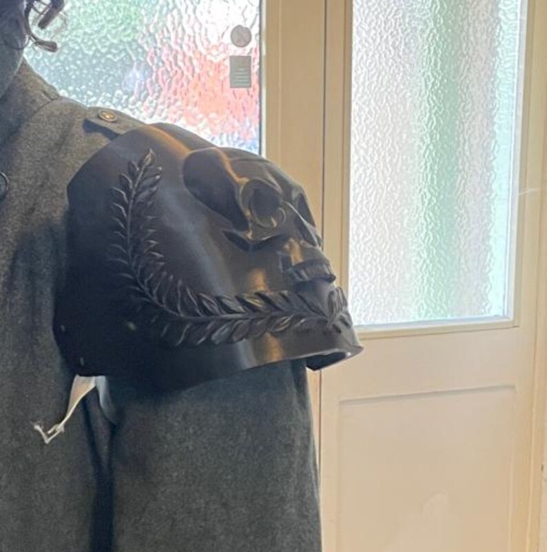 May include: A black shoulder pauldron with a skull design and laurel leaf accents. The pauldron is worn on a gray coat, showcasing the intricate details of the skull and leaf carvings. The design is a gothic or fantasy theme.
