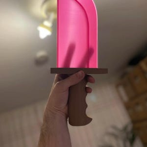 May include: A pink sword with a brown handle and crossguard. The blade is a vibrant pink, and the handle is designed for a comfortable grip. The sword is held in a hand, suggesting it is a prop or toy.