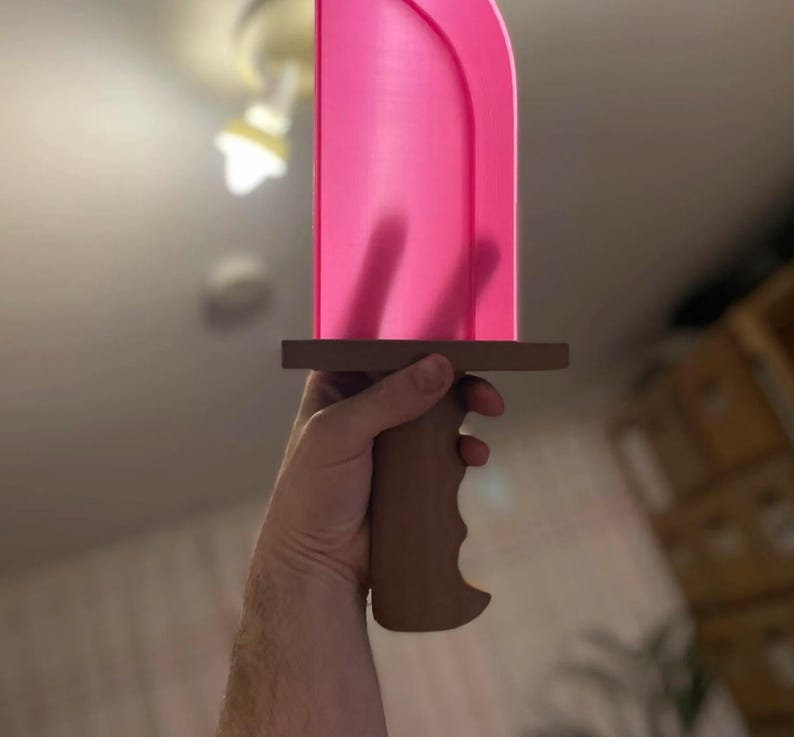 May include: A pink sword with a brown handle and crossguard. The blade is a vibrant pink, and the handle is designed for a comfortable grip. The sword is held in a hand, suggesting it is a prop or toy.