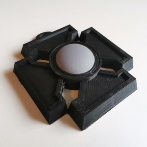May include: A black, geometric, 3D-printed object with a central grey, circular component. The object has a symmetrical design with four outward-facing, angular sections. The grey centre has a textured surface, and the overall design is modern and abstract.